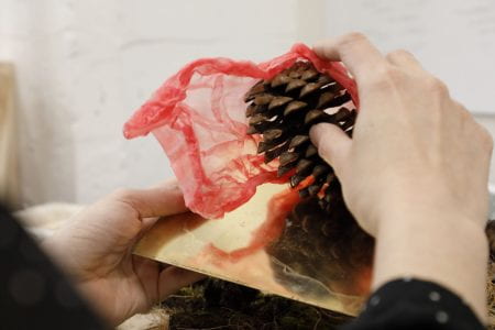 Image of an artist experimenting with materials: a pine cone, pink rubber latex and golden metal square