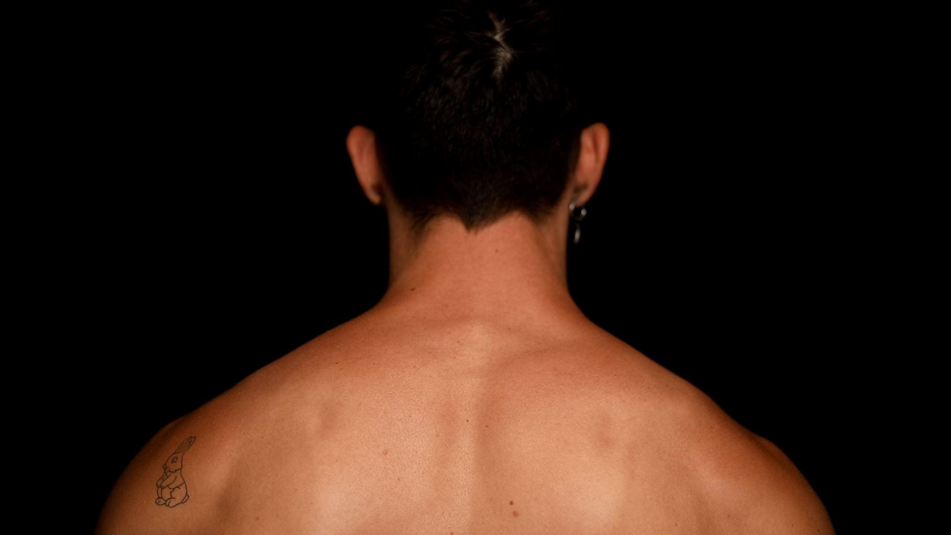 Back of a person's head with shoulder and neck showing. They have defined musculature and a tattoo of a rabbit.