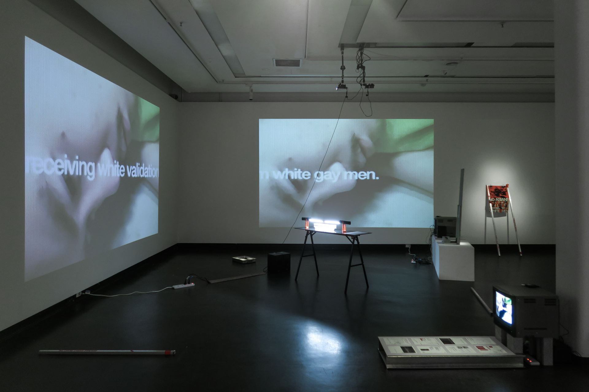 an image of the 'miscegenasian' project in a gallery, containing two projections, a trestle table, two videos on CRT televisions, steel beams on the ground, and a sign that reads 'No Asians Allowed Unless Accompanied By White Men'.