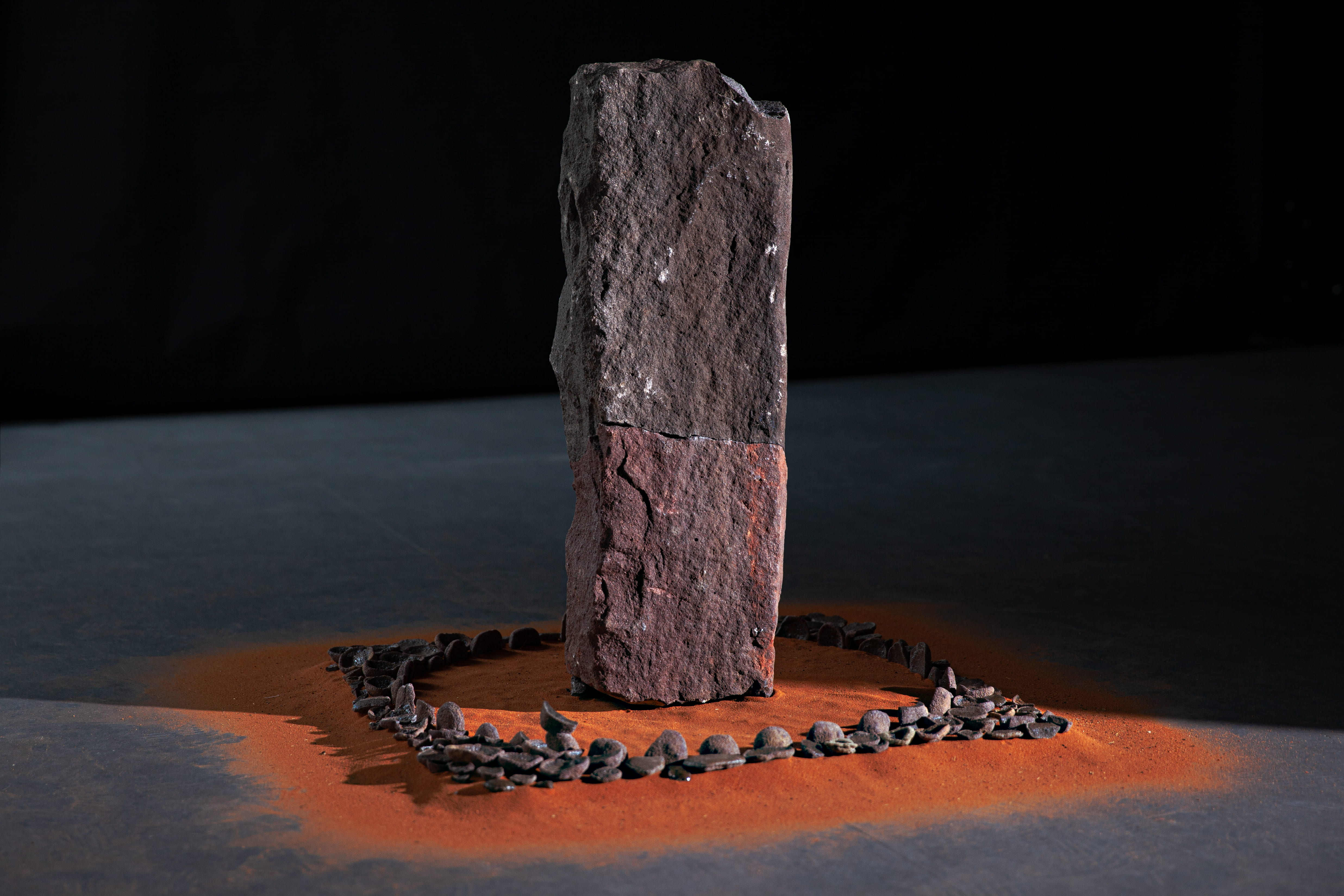 A photograph of a sculpture. A pillar of stone stands upright on a bed of red sand, surrounded by a circle of small rocks. The bottom third of the pillar is a reddish purple colour, while the rest is a natural blue-grey. 