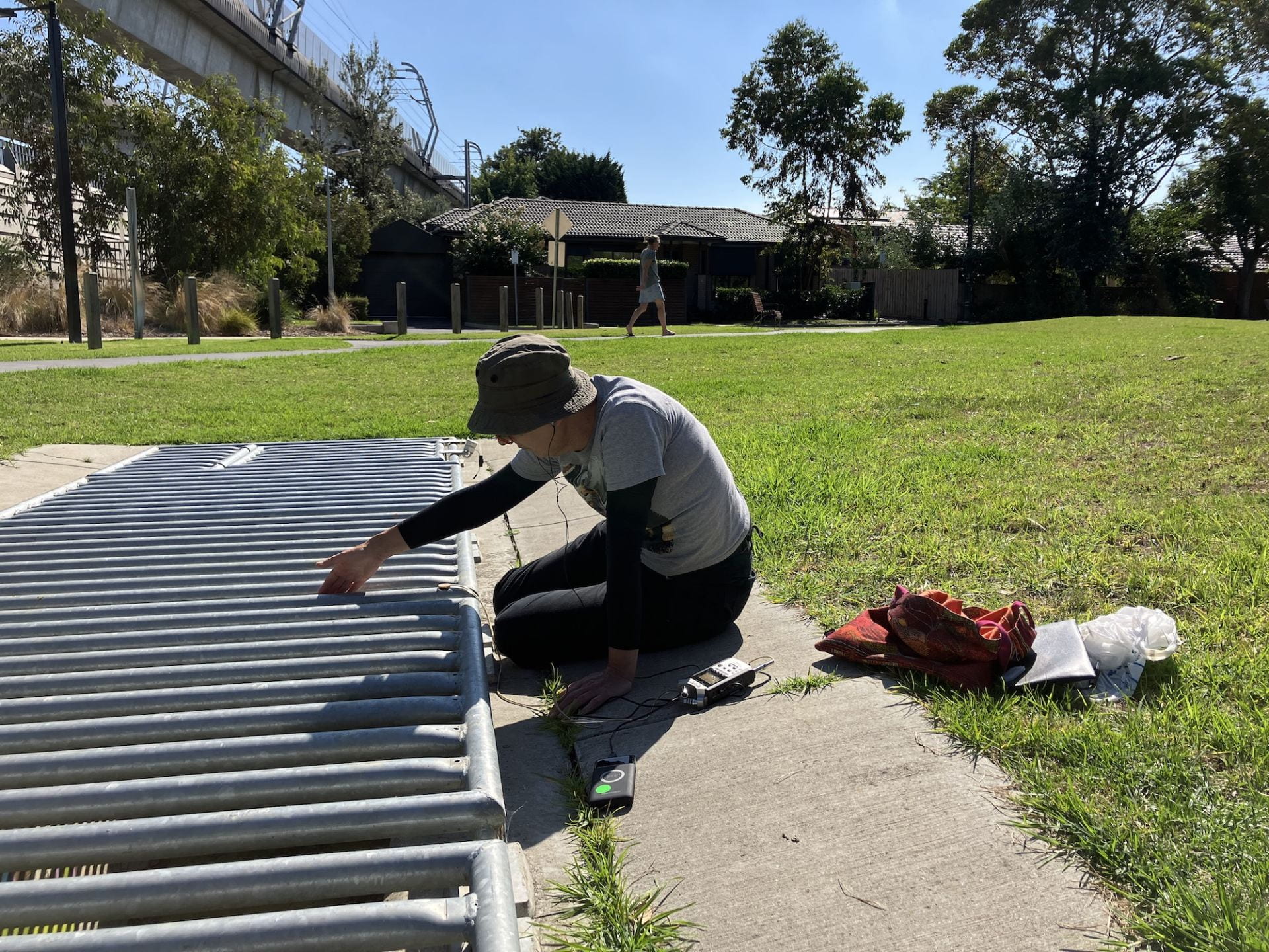 This image is of Vivienne Tate taking field recordings for the Murrumbeena Station Precinct project. She is using a contact mic to record the sounds of the metal drain grill.