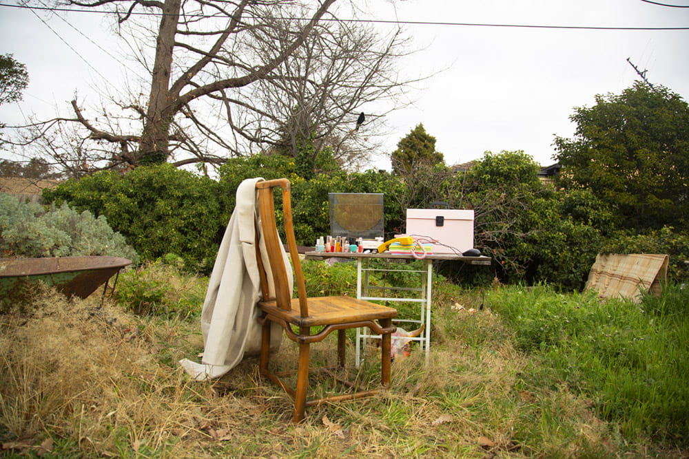 In an overgrown backyard sits a table and chair. The table is full of colourful objects that could be found in a work desk.