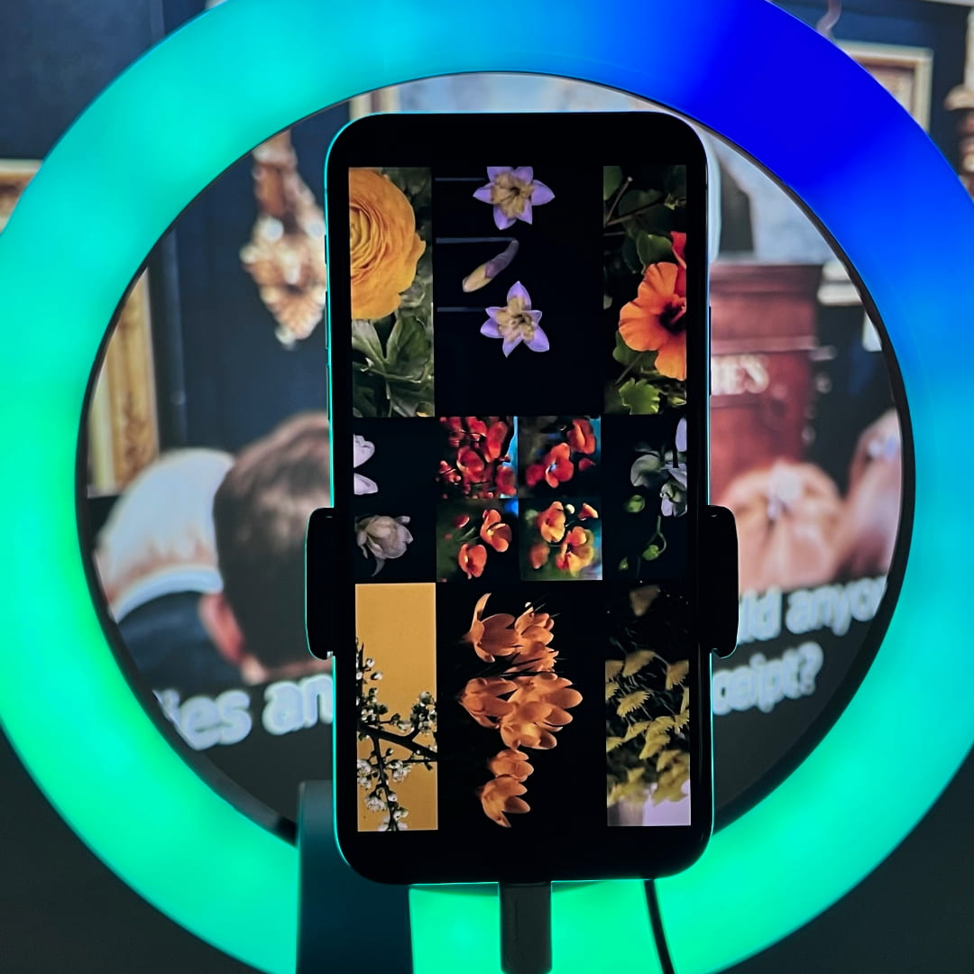 Photograph of a phone displayed on a green and blue ringlight. The phone shows an image of AI generated flowers next to real flowers. Behind the phone, there is a video projection of a Christie's auction.