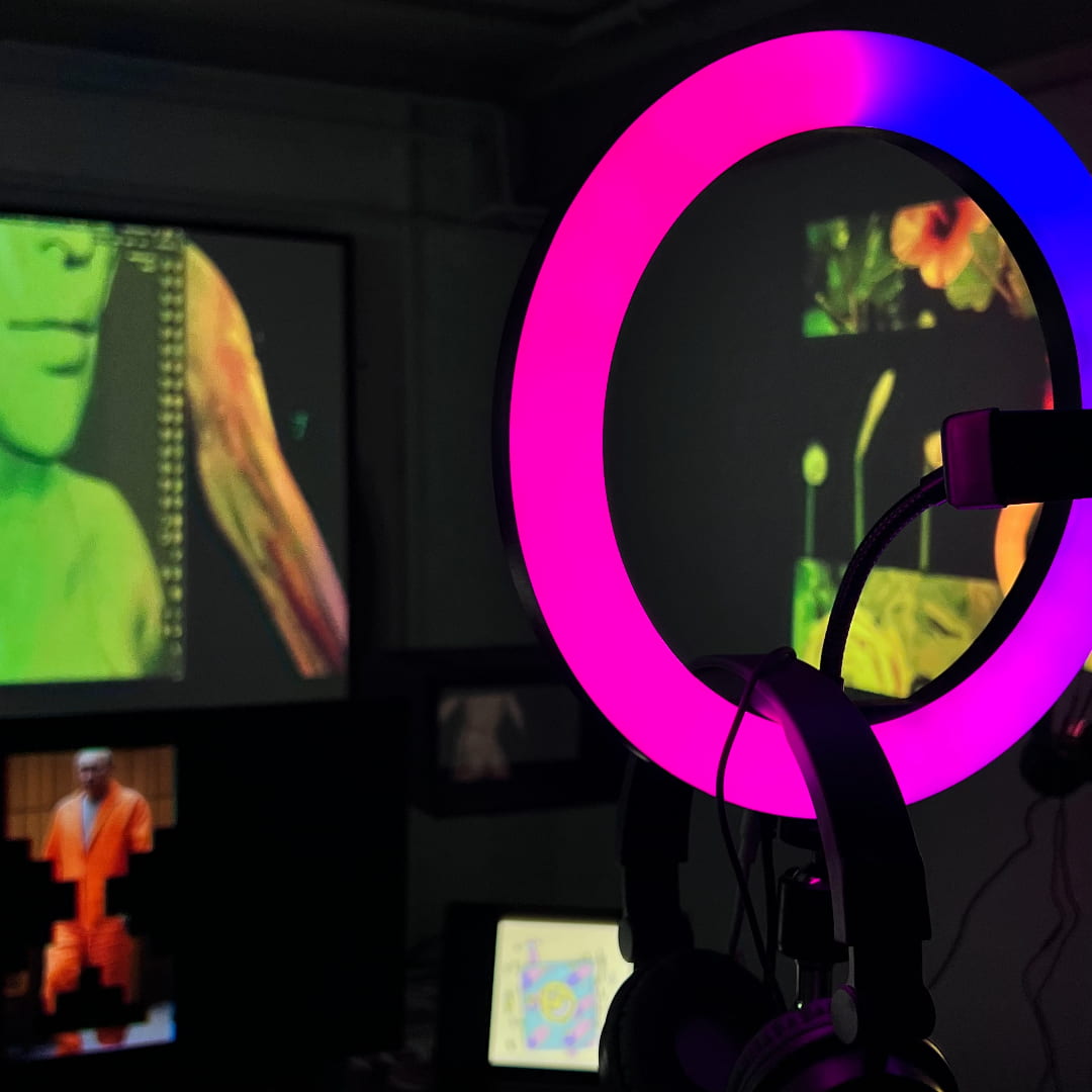 Photograph of a dark room with multiple video projections and TV screens. In the foreground, there is a pink and blue ringlight. Behind it, there are blurred images of Putin, AI generated flowers, and 3D modelled people.