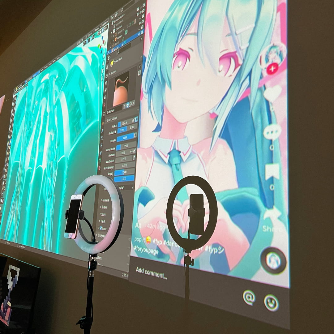 Photograph of a low light room with a phone displayed on a ringlight. Behind it, there is a large video projection of Hatsune Miku and 3D modelled jellyfish displayed on the Blender interface.