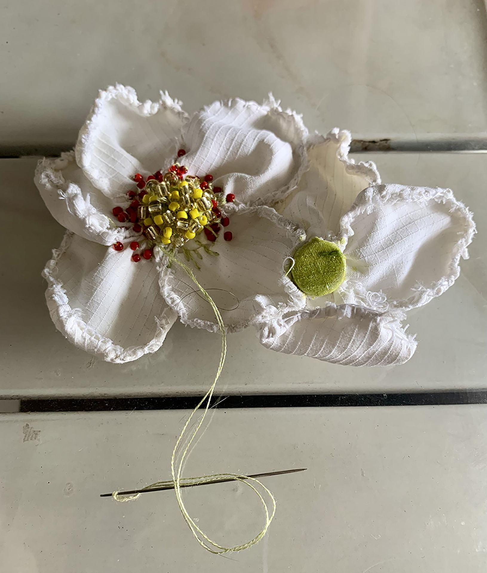 Textile flowers under construction with a hand-sewing needle and thread, embellished with beads and resting on a white slatted table top.
