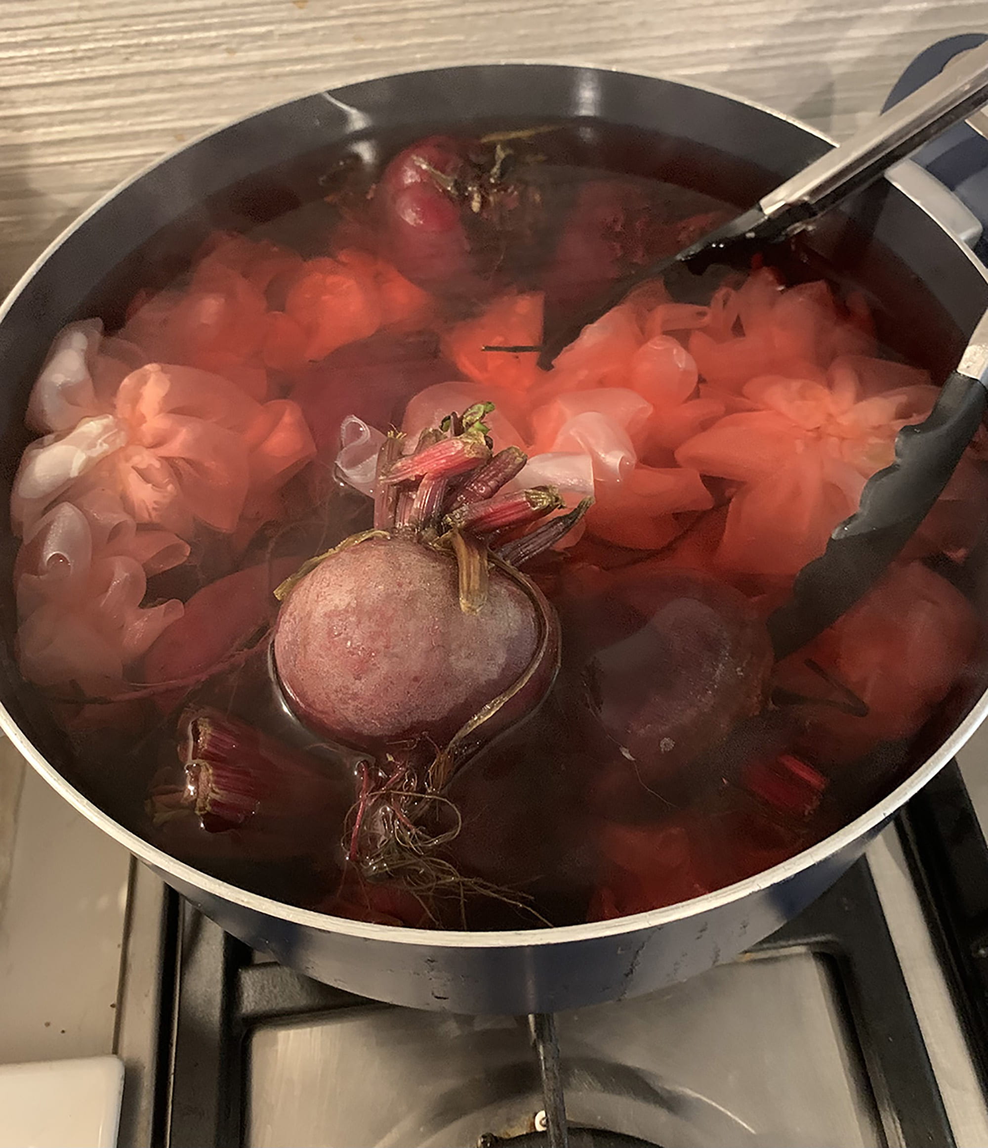 Beetroot and voile constructs in a cooking pot on a stovetop with silicone-tipped metal tongs.