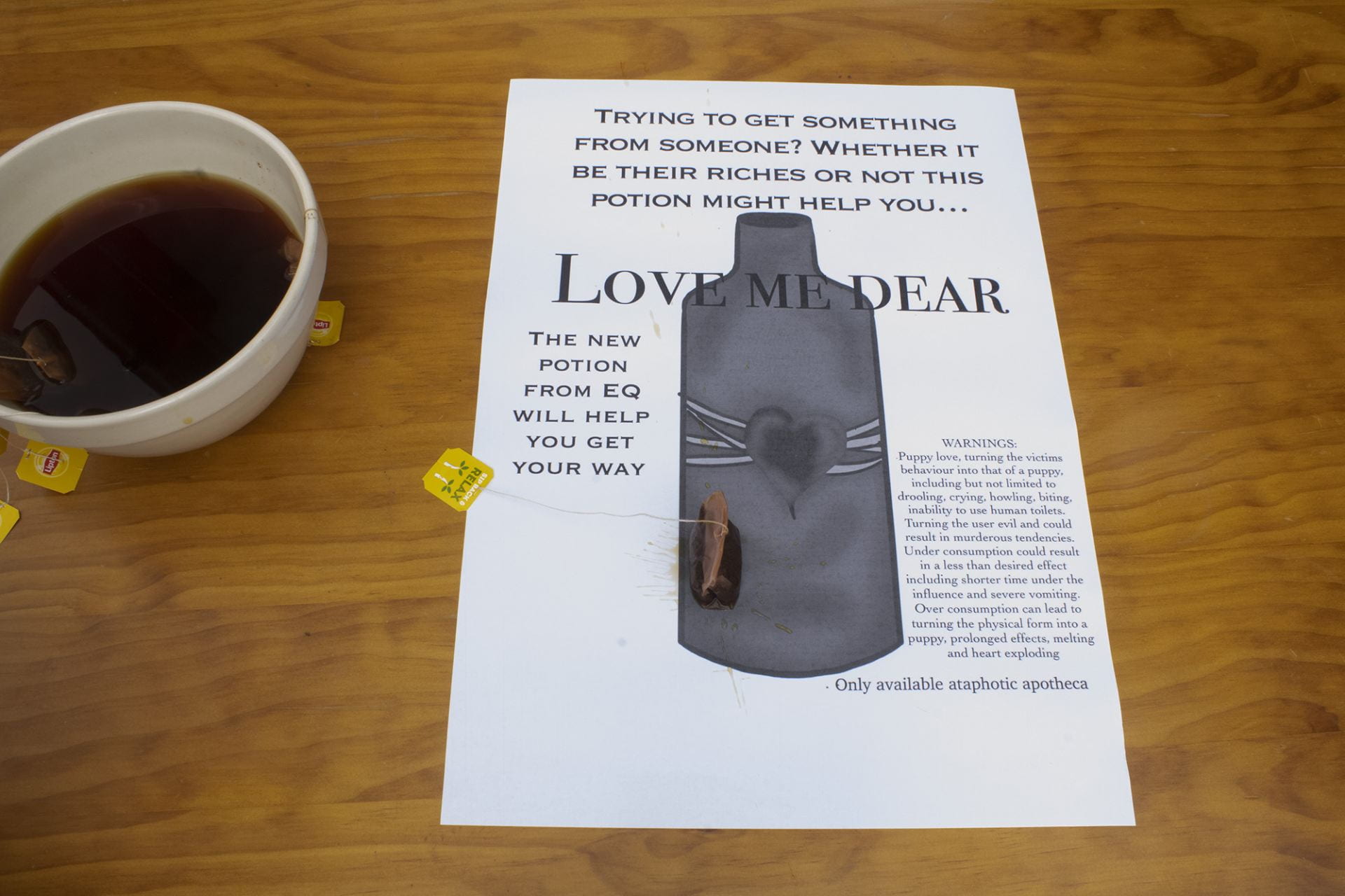 A3 poster with an image of a bottle and text saying Love Me Dear. A bowl of tea on the left and a tea bag on the poster.