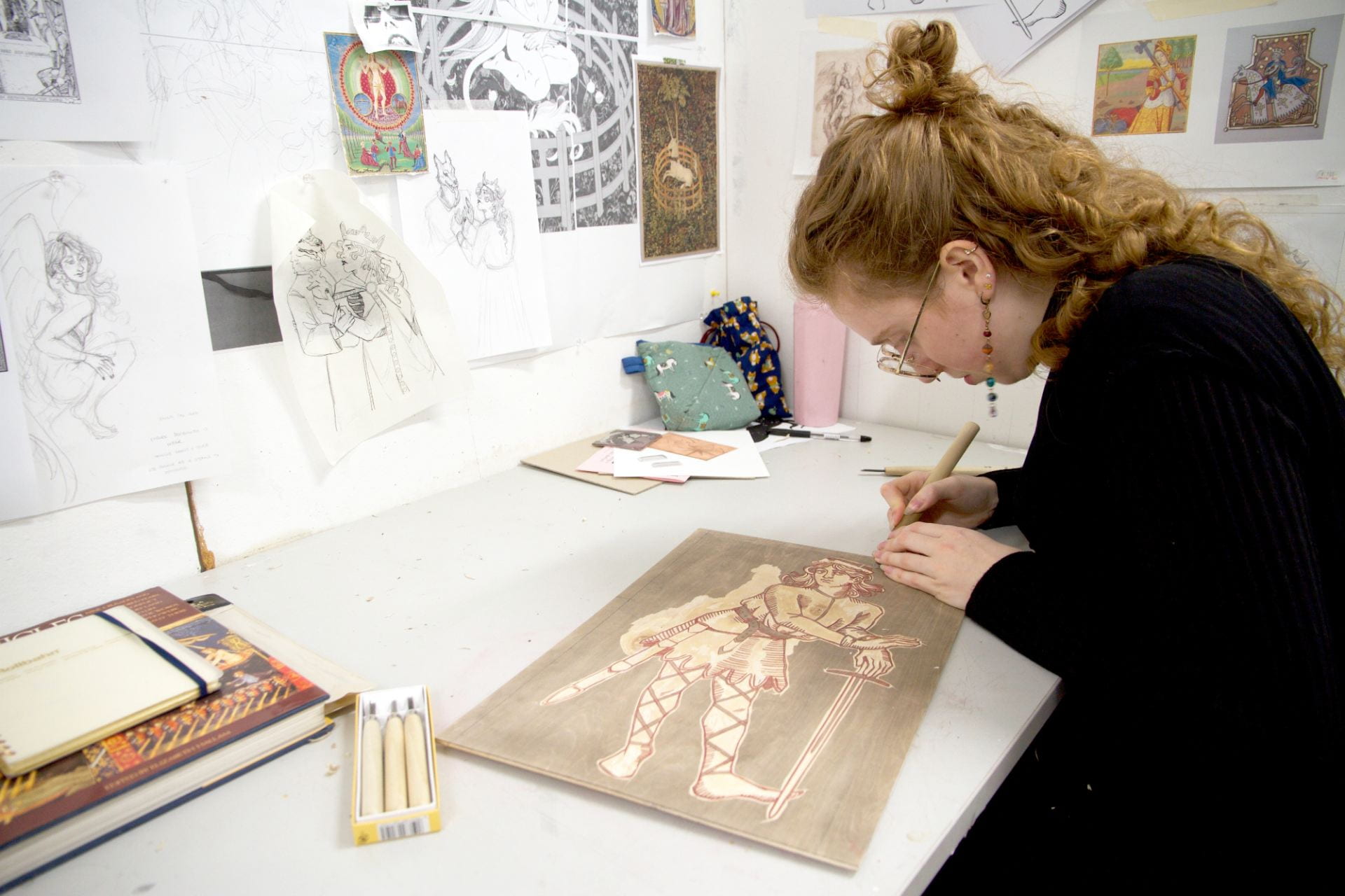 artist sat at desk working on woodcut, wall behind is covered in images.