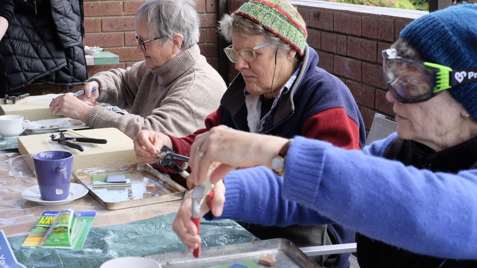 Three older woman sit at a table cutting tiles with tools for a mosaic. They wear glasses and goggles.