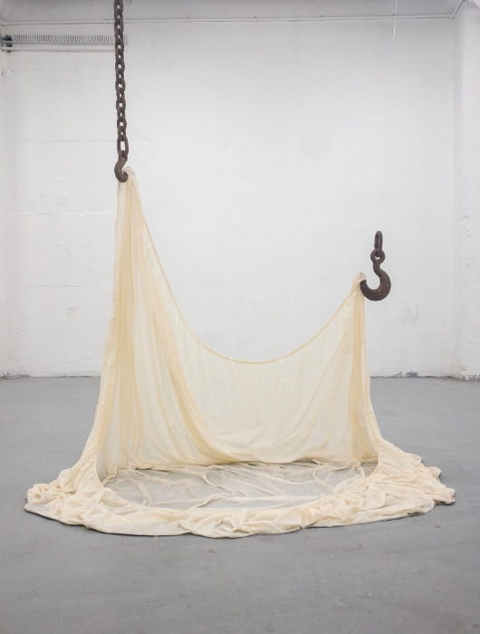 A rusty chain hangs down and so does a large rusty hook.  Between the two a white sheet is strung. 