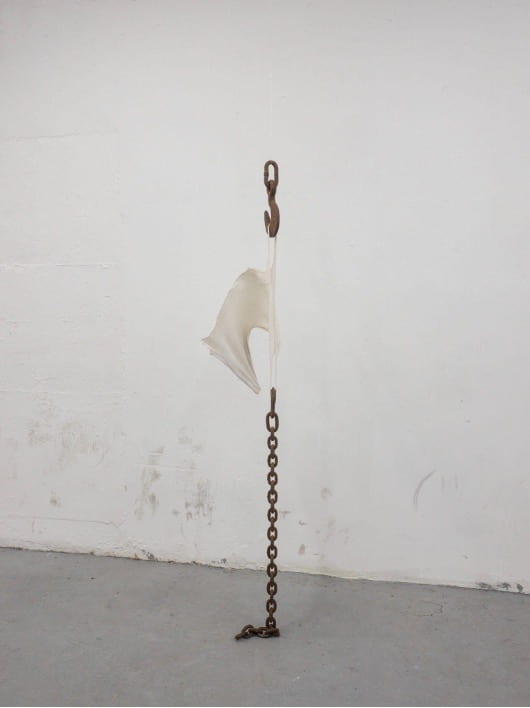A rusty hook hangs from the ceiling. A piece of torn white fabric hangs from. Attached to the fabric there is a rusty chain the weight of which is tearing the fabric. 