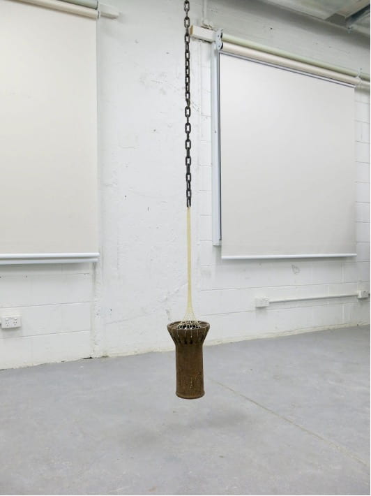A large and heavy section of rusty, metal pipe hangs from the ceiling. It is held in place by a thin web of crochet. 