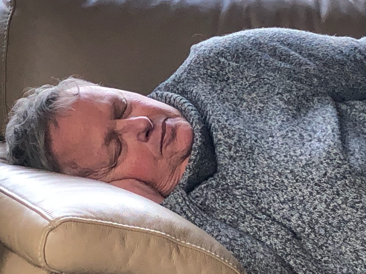 An older woman in a grey jumper sleeps on a couch