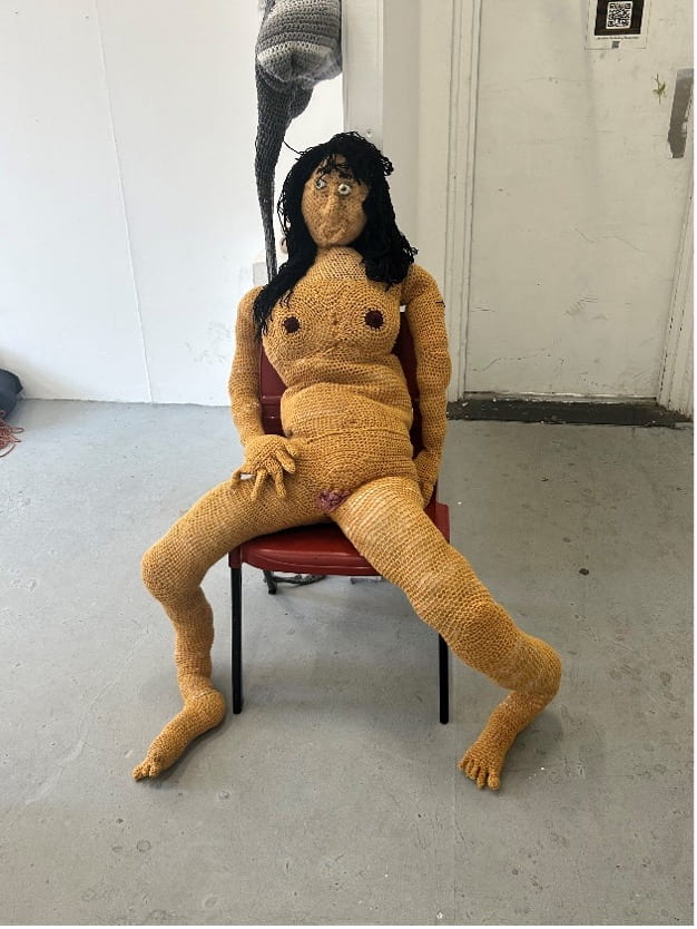 A crochet, naked woman lounges on a maroon chair. 