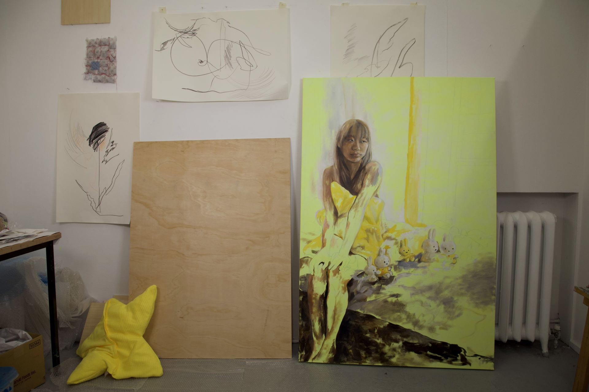 A view of the artists' studio. In the centre of the image there is a yellow painting of girl in wood next to a large panel of wood.