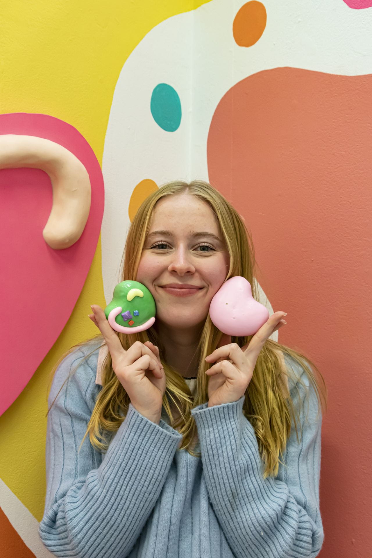 Artist posing with two small heart sculptures near face, with mural behind.
