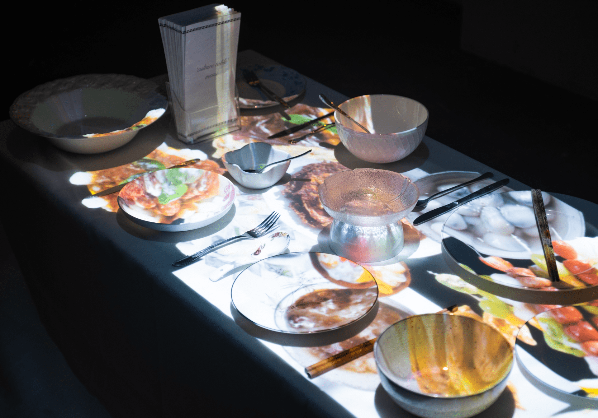 Photograph of a dinner table installation artwork. There are images of colourful multicultural food projected onto the table onto white plates and glassware.