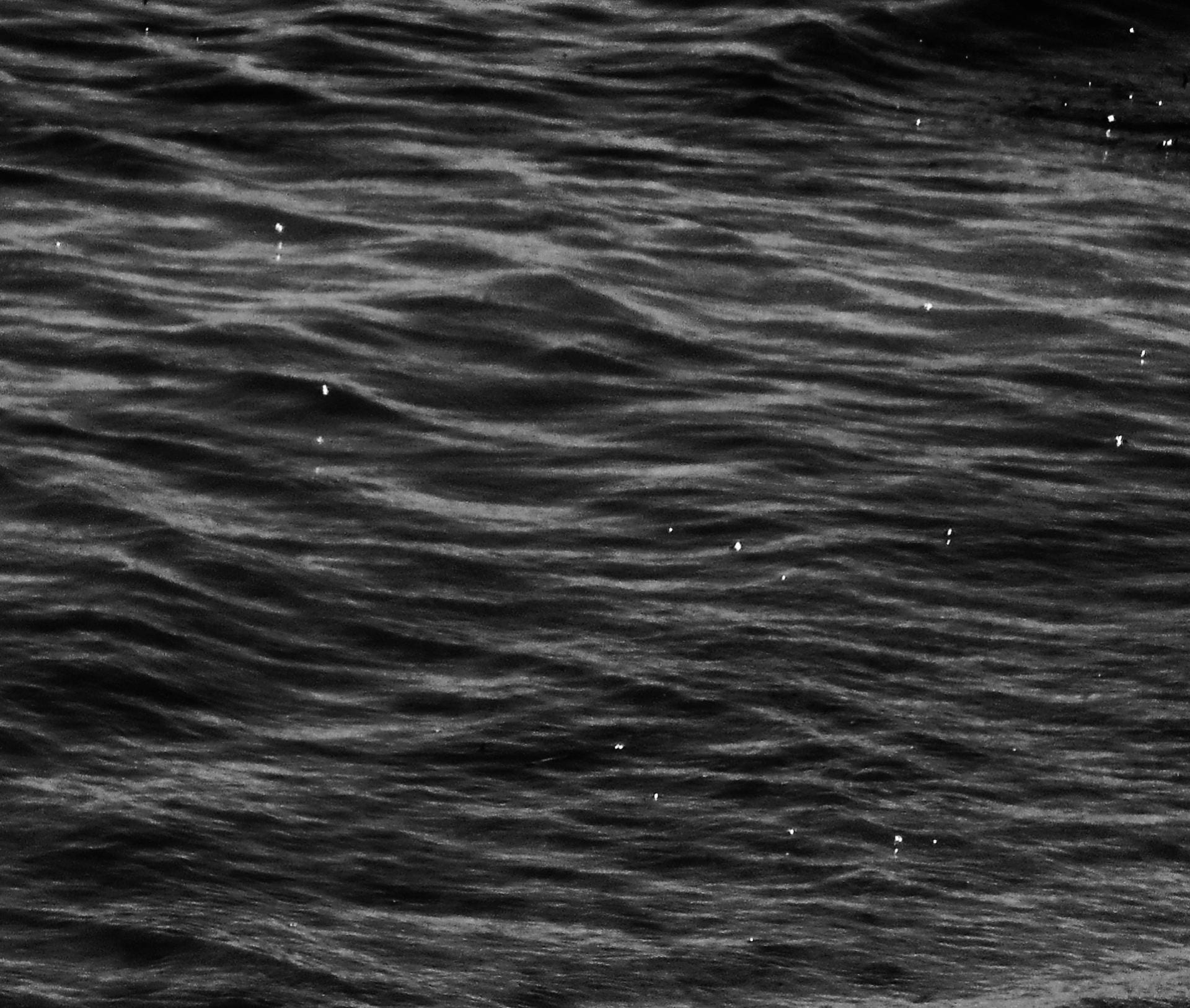 black and white image of the surface of water. it is cropped in close, no waves visible. There are small white dots, specks of light reflecting on the spray of the sea.