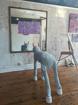 A light grey animal with four legs, white hooves and red demon horns stands looking in a mirror. The mirror has a short story written on it. 