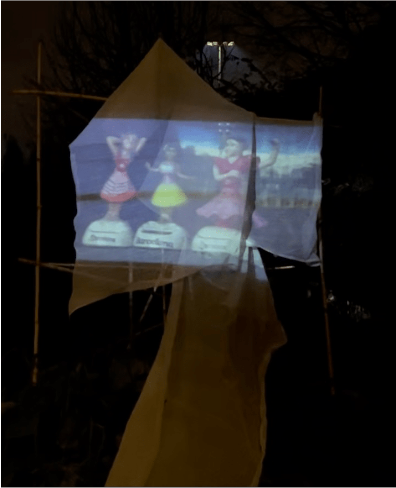 A projection on a sheet in a garden depicts a photo of a car dashboard. Three kitsch bobble-figurines of women sit on the dashboard with a name of a different city below them. 