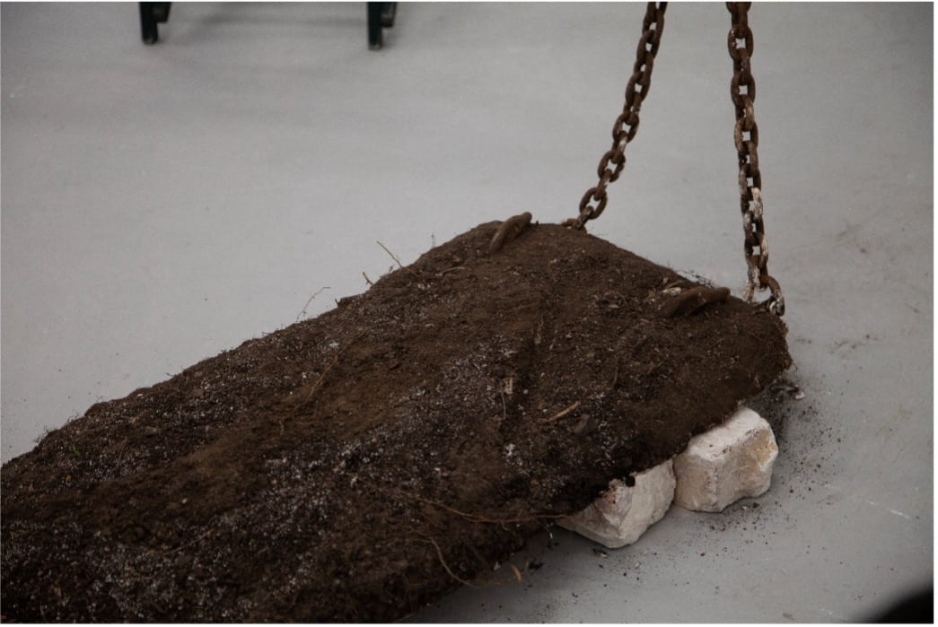 A slab of soil sits on a bed of plaster balancing on bricks. A large chain elevates the bed above the floor. 